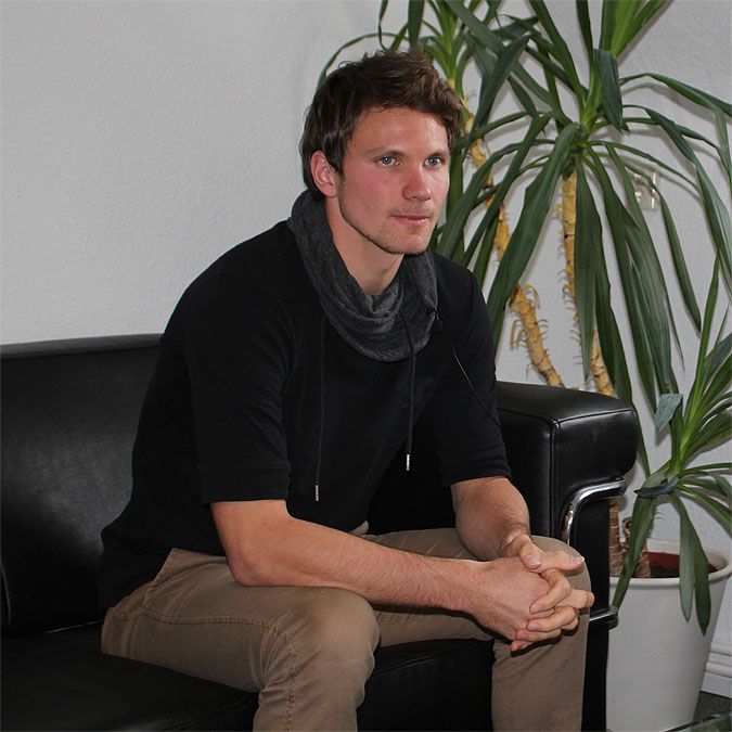 Ein junger Mann sitzt auf einer schwarzen Couch, trägt einen schwarzen Pullover und hat einen grauen Schal um den Hals.