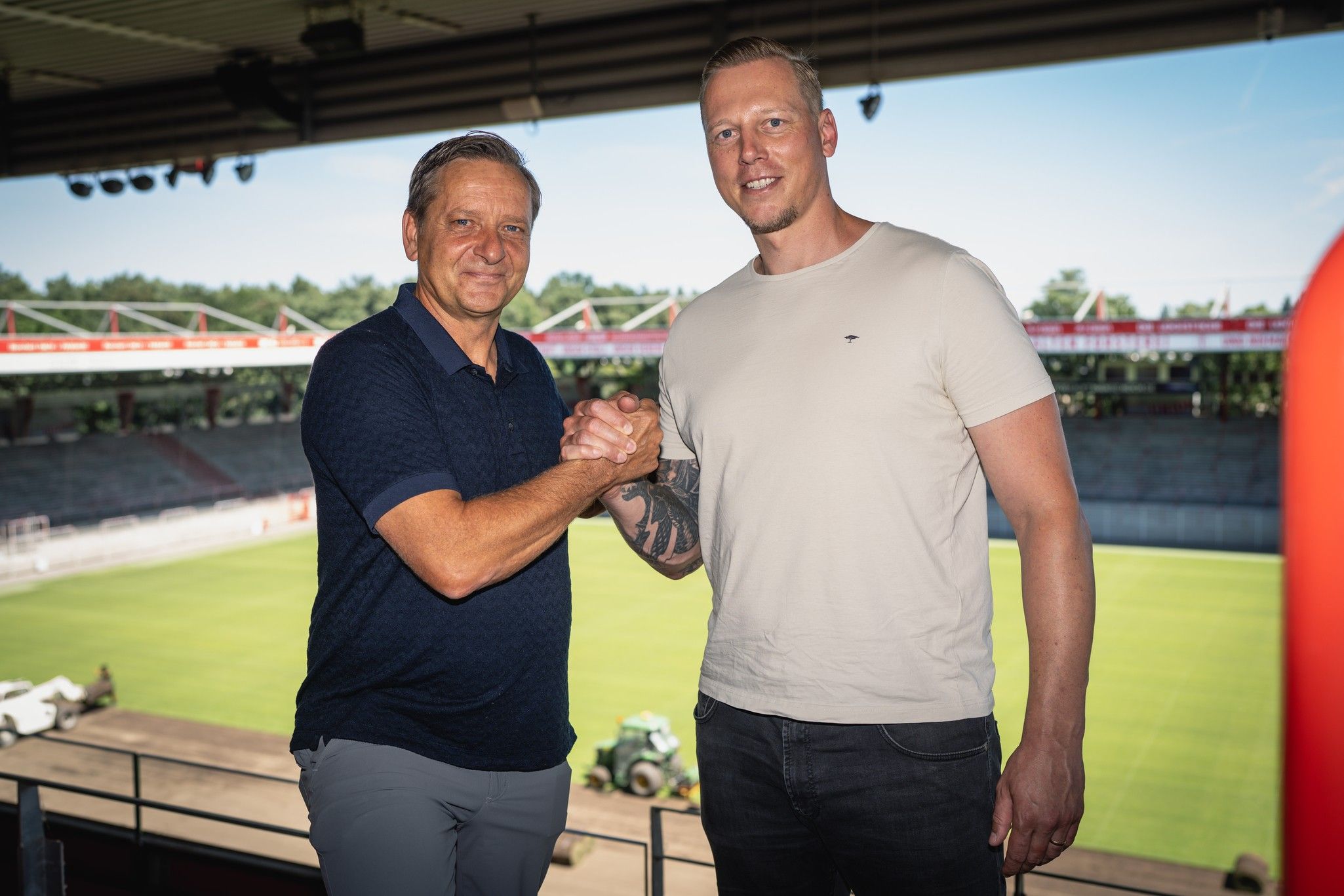 Zwei Männer schütteln sich in einem Stadion die Hände, im Hintergrund ist das Spielfeld sichtbar.