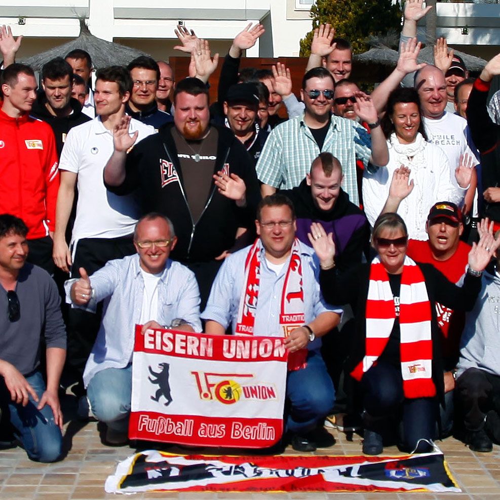 Eine Gruppe von Menschen steht lächelnd im Freien, einige tragen Fanartikel und halten ein Banner von "Eisern Union".