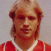 Ein junger Mann mit blonden Haaren trägt ein rotes Sporttrikot und blickt in die Kamera.