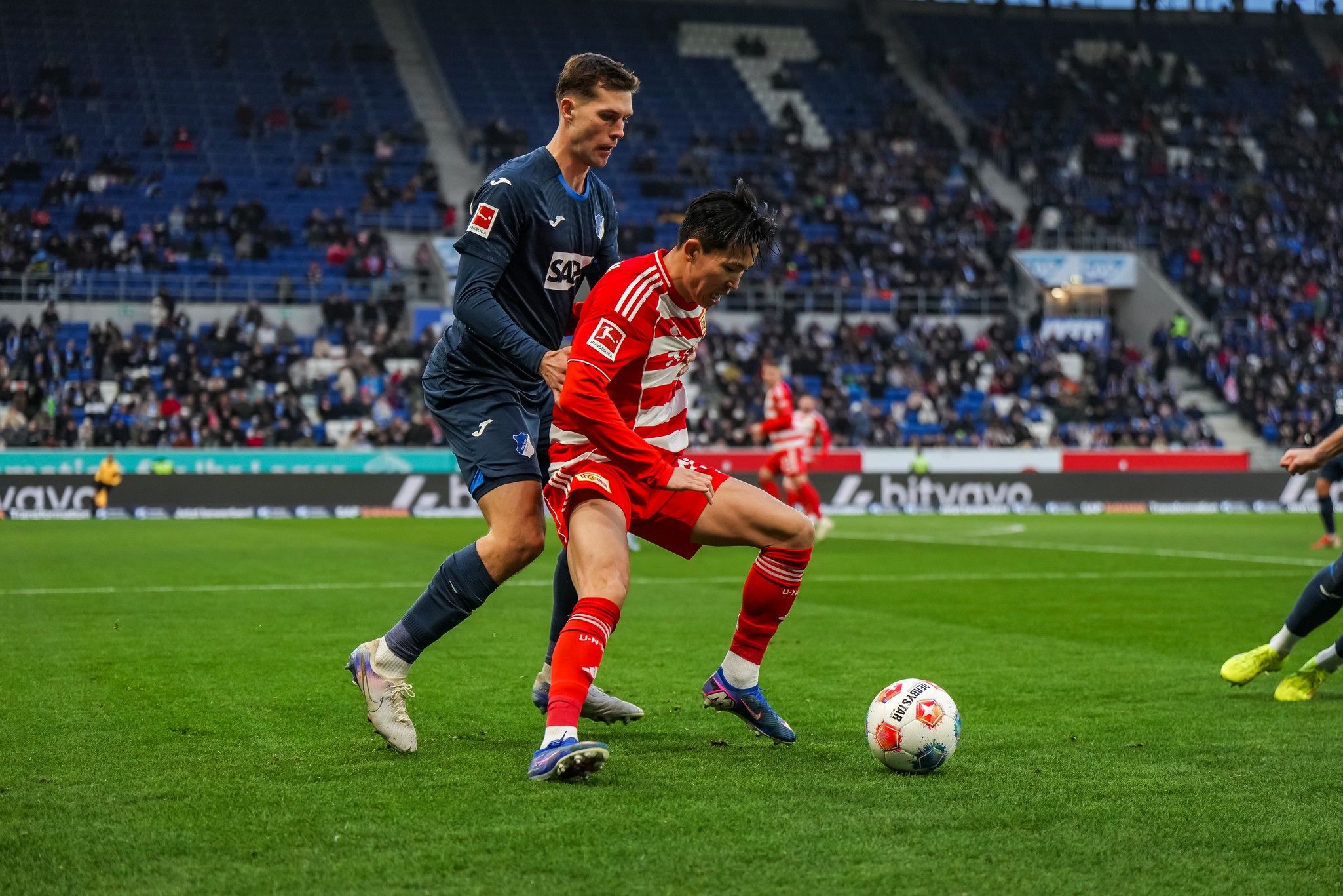 Zwei Fußballspieler kämpfen um den Ball auf einem Spielfeld, während Zuschauer im Hintergrund sitzen.