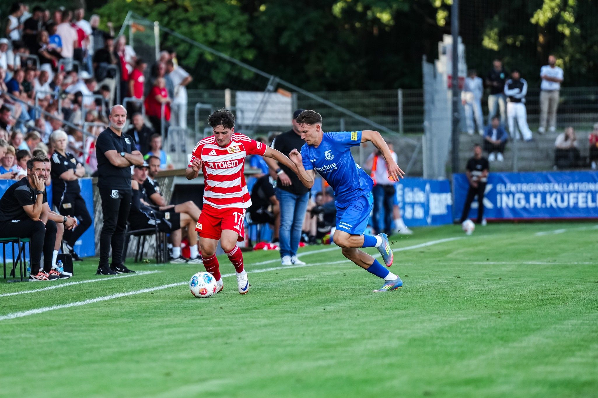 Ein Fußballspieler in roten Trikots dribbelt den Ball, während ein gegnerischer Spieler in Blau ihn verfolgt. Zuschauer sind im Hintergrund.