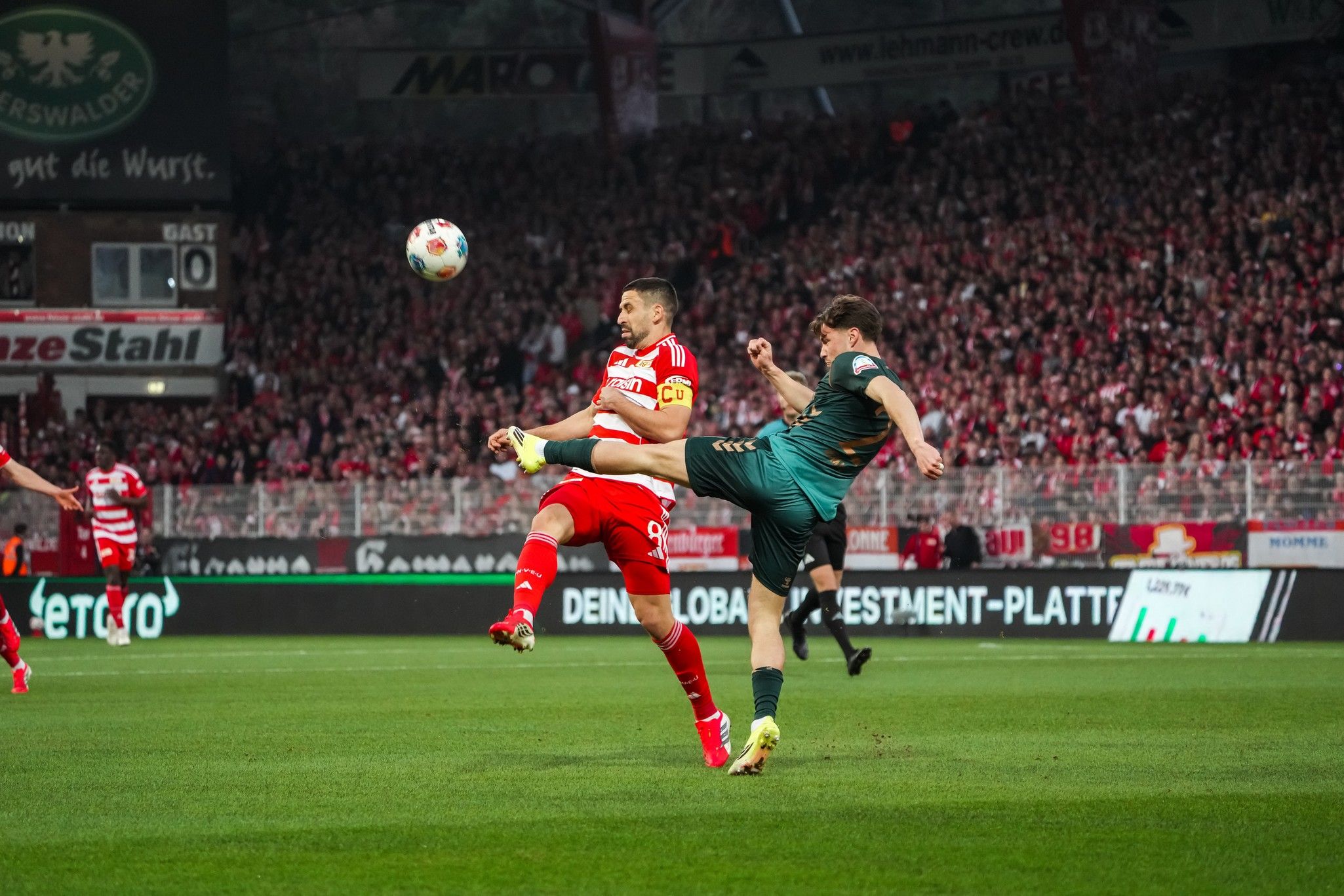 Ein Fußballspieler in roter Uniform hebt das Bein, während ein Spieler in grünem Trikot ihm entgegenkommt. Stadion mit Zuschauern im Hintergrund.