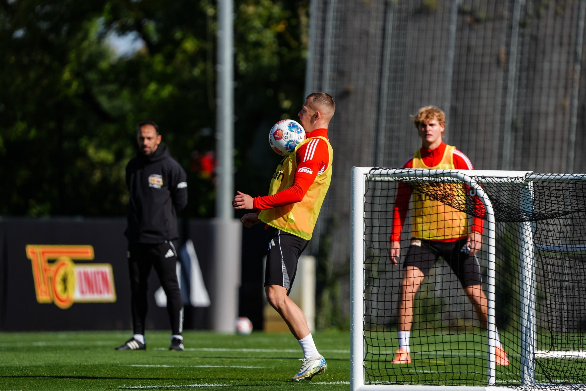 Die Szene zeigt zwei Fußballspieler im Training, während ein dritter Spieler im Hintergrund zuschaut.