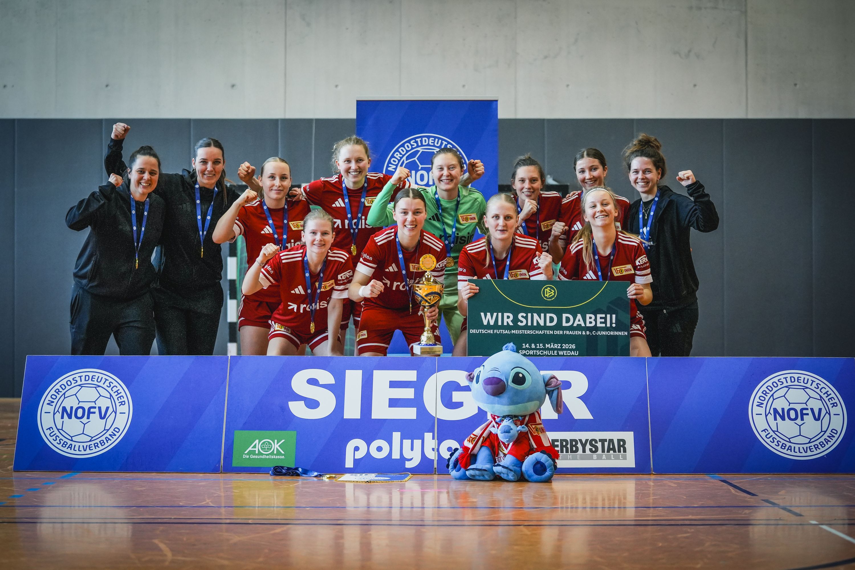 Frauenmannschaft zeigt ihren Sieg mit Pokal, jubelnd in Sportoutfits, vor einem Banner und einem Plüschtier.