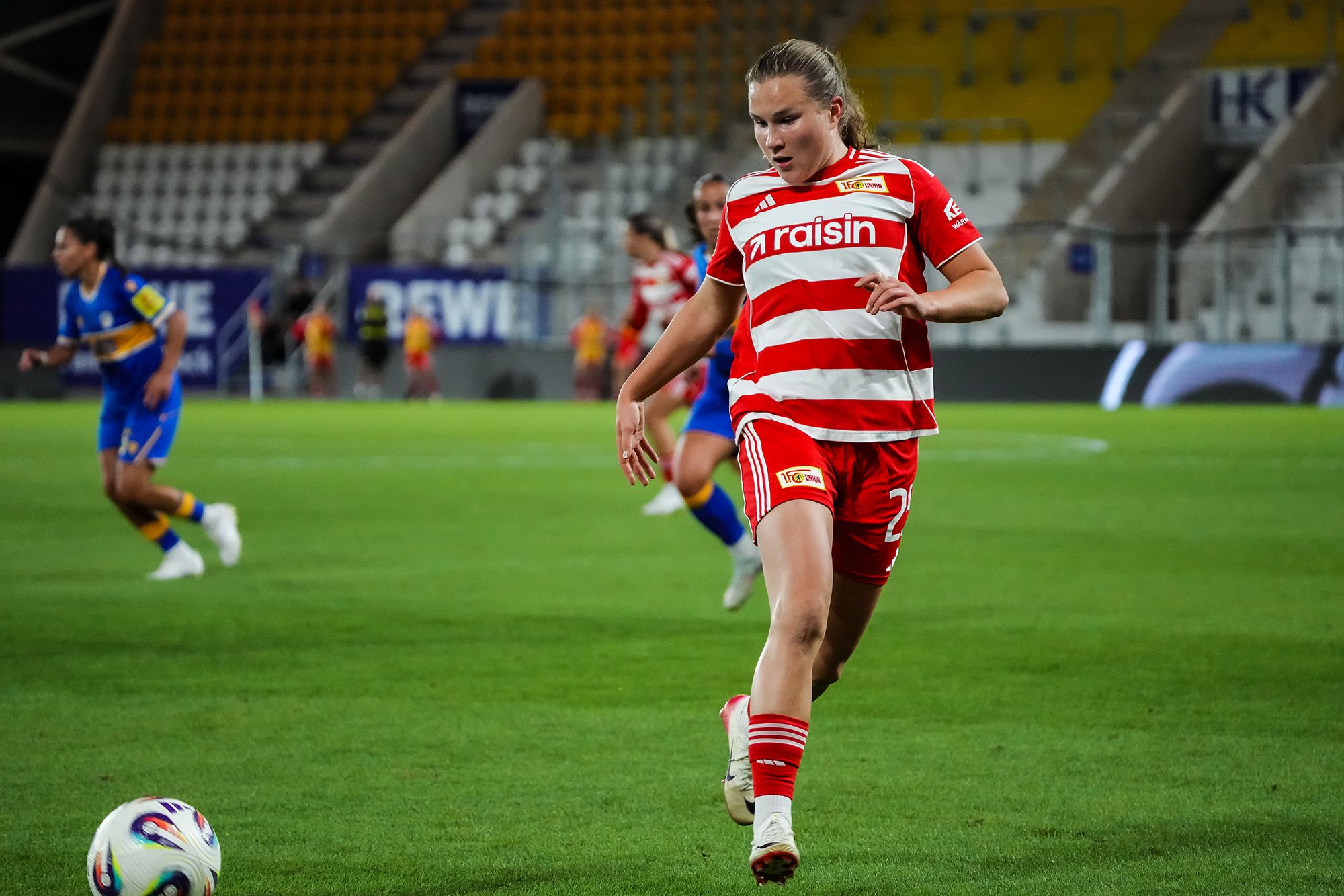 Spielerin in gestreiftem Trikot dribbelt den Fußball auf dem Feld während eines Spiels. Stadionatmosphäre im Hintergrund.