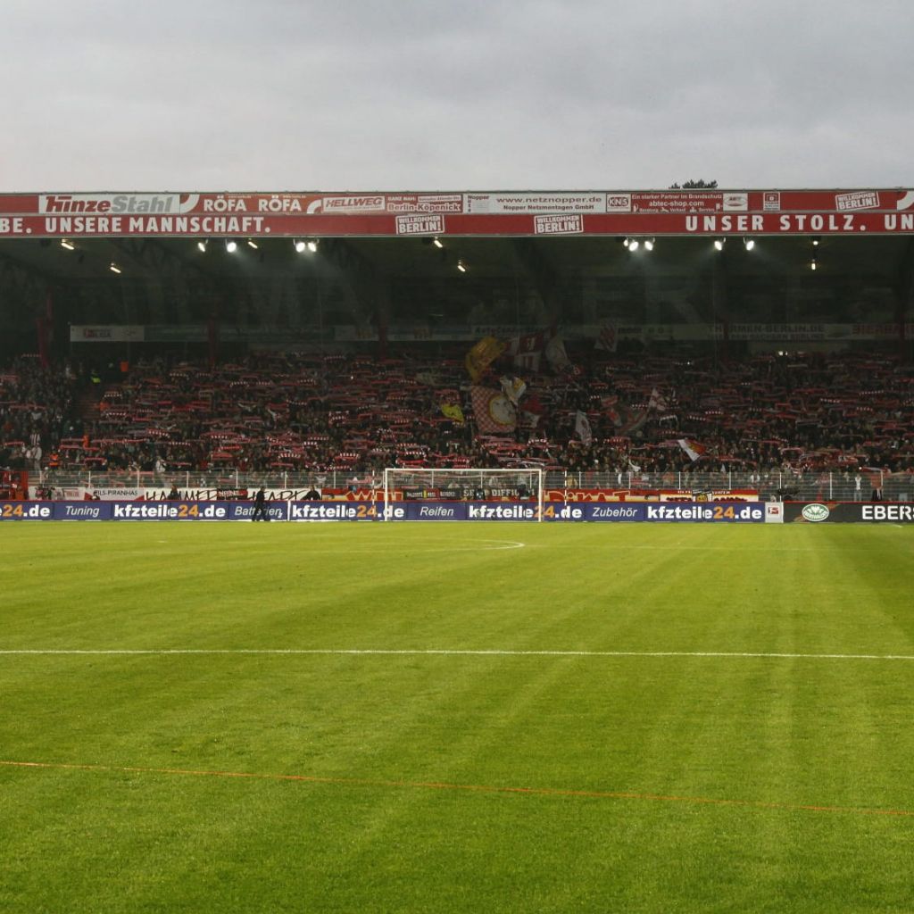Fußballstadion mit einem voll besetzten Fanblock und einem gepflegten Rasenfeld im Vordergrund. Wolkiger Himmel.