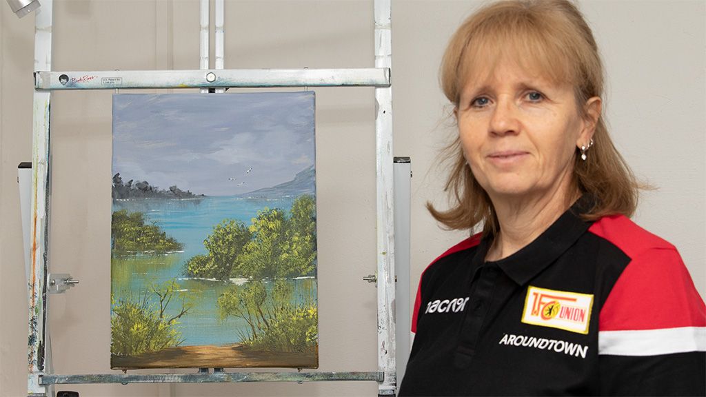 Eine Frau steht neben einem Gemälde einer Landschaft mit Wasser und Bäumen, das auf einem Staffelei hängt.