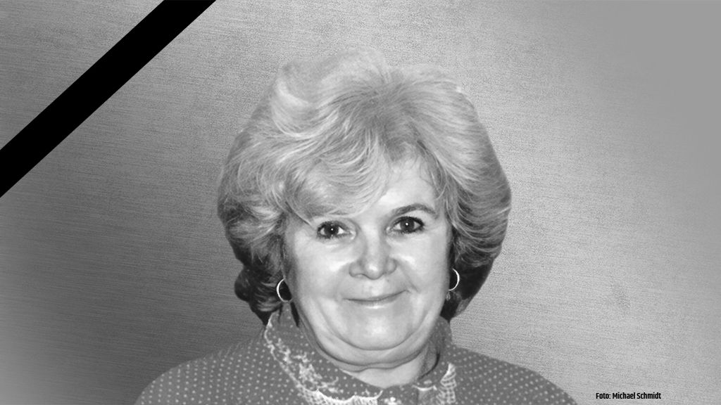 Porträt einer älteren Frau mit lockigem Haar, die in die Kamera lächelt, vor einem grauen Hintergrund und einem schwarzen Trauerrand.