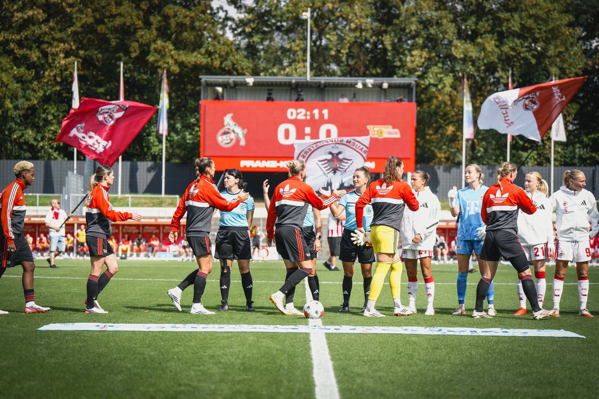Zwei Fußballteams begrüßen sich vor einem Stadion, im Hintergrund steht die Spielstandsanzeige bei 0:0.