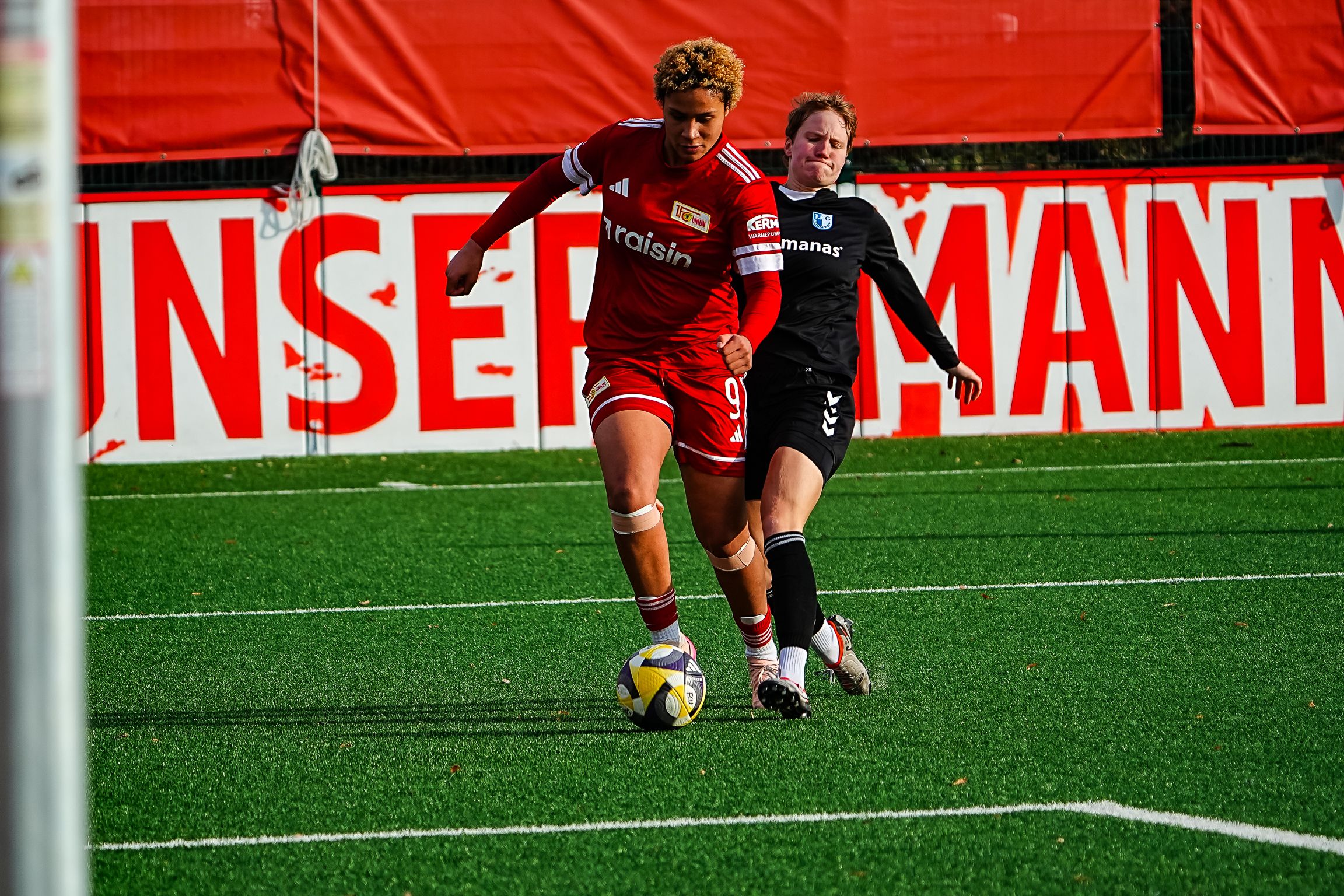 Eine Spielerin in roter Uniform dribbelt den Ball, während eine gegnerische Spielerin in Schwarz versucht, sie zu stoppen.