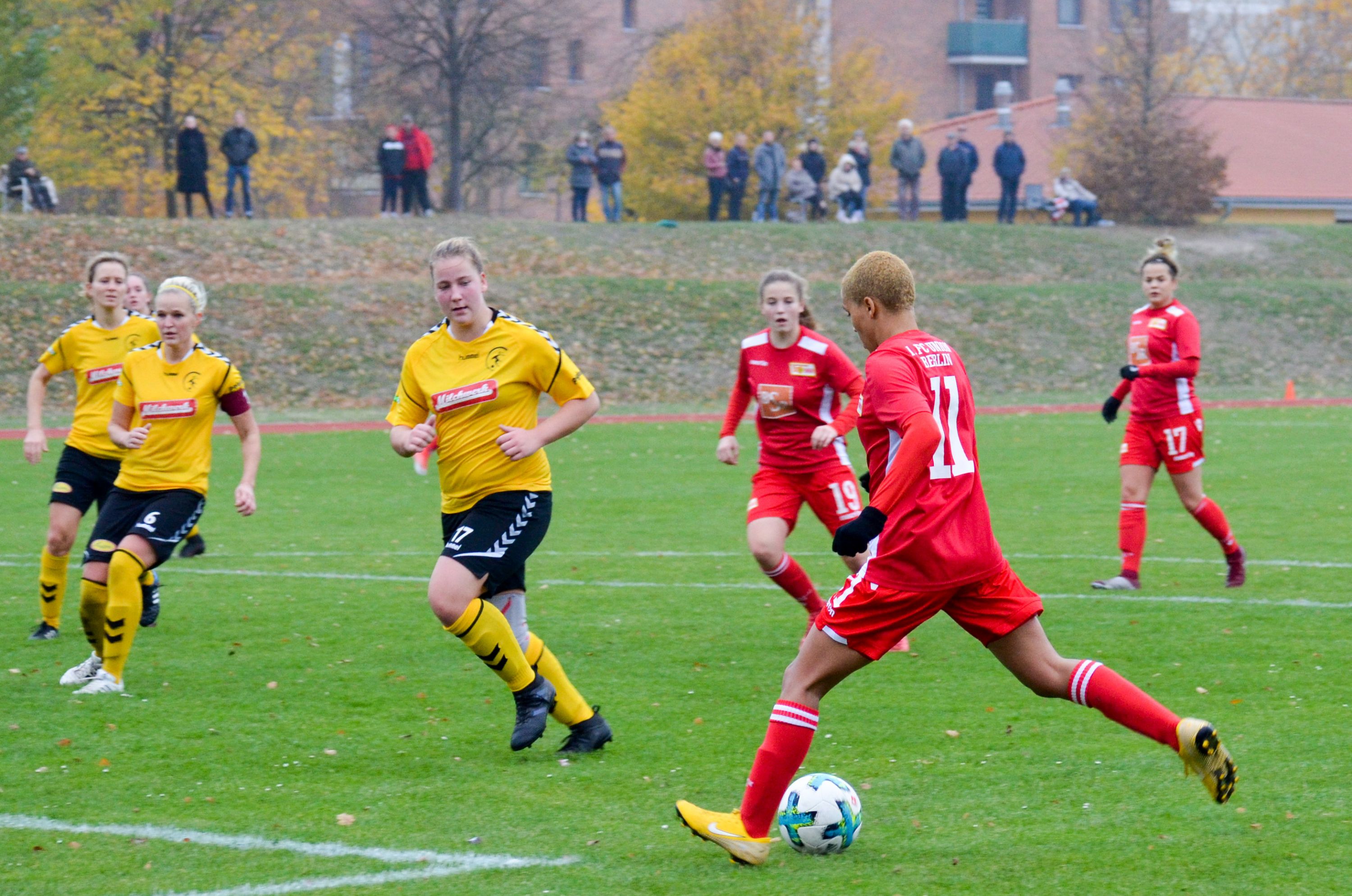 Spielszene eines Fußballs mit Spielerinnen in roten und gelben Trikots, die einem Ball auf einem grünen Rasenfeld nachjagen.