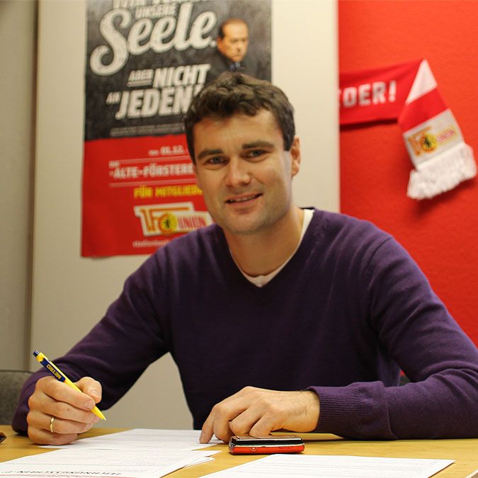 Ein Mann sitzt an einem Tisch, schreibt Notizen und blickt zur Kamera, im Hintergrund ein Plakat und eine Fahne.