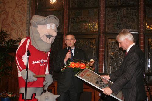 Ein Maskottchen in Sportkleidung steht neben einem Redner, während ein Mann ein Bild überreicht. Blumenarrangement im Vordergrund.