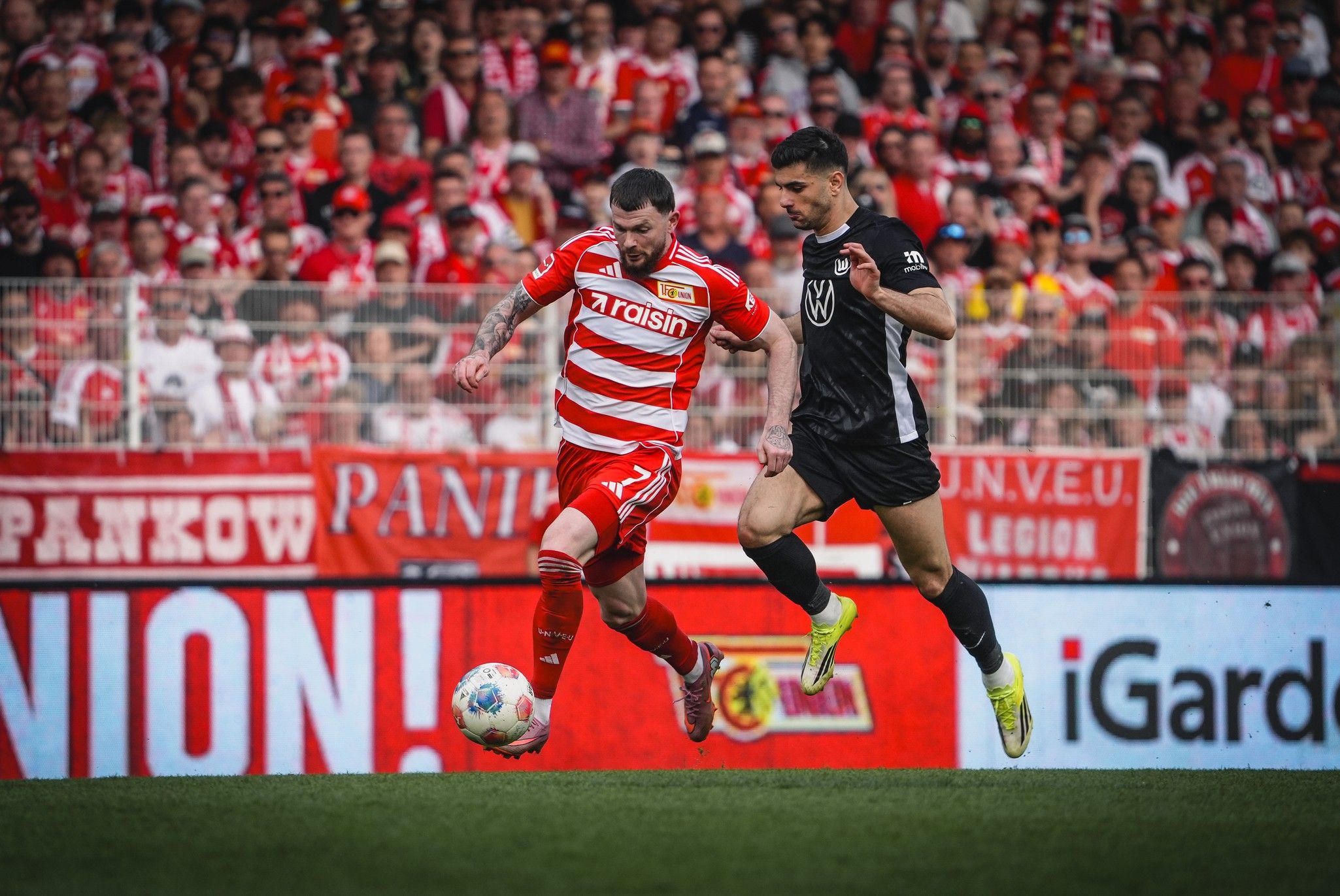 Spielszene mit zwei Fußballspielern, einer in rot-weiß gestreiftem Trikot, der andere in schwarzer Kleidung, während sie um den Ball kämpfen.