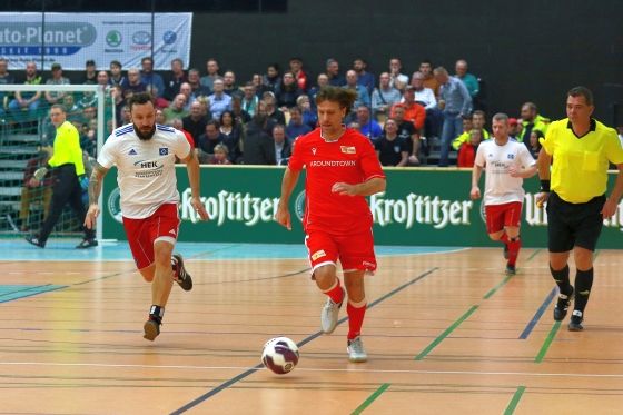 Zwei Fußballspieler im Wettkampf, einer in rotem Trikot, der andere in weißem Trikot, mit Zuschauern im Hintergrund.