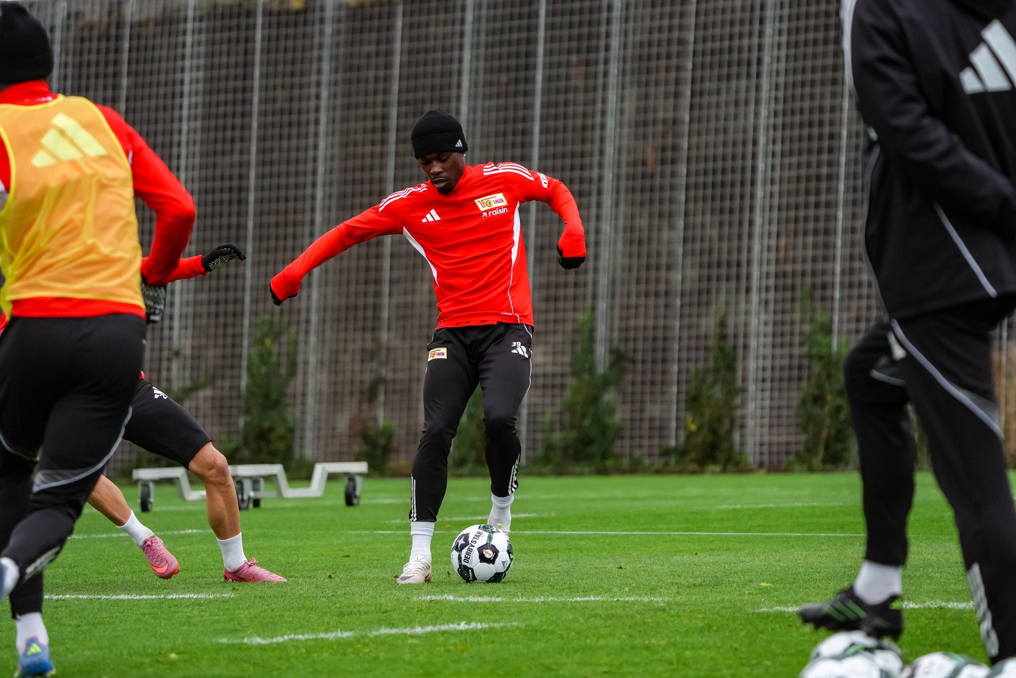 Fußballspieler im Training, der einen Ball auf dem Rasen dribbelt, umgeben von weiteren Spielern in Trainingskleidung.