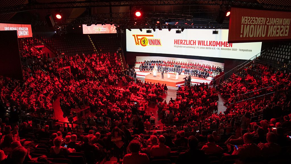A large hall with a crowd attending an event, featuring a stage and large screens in the background.