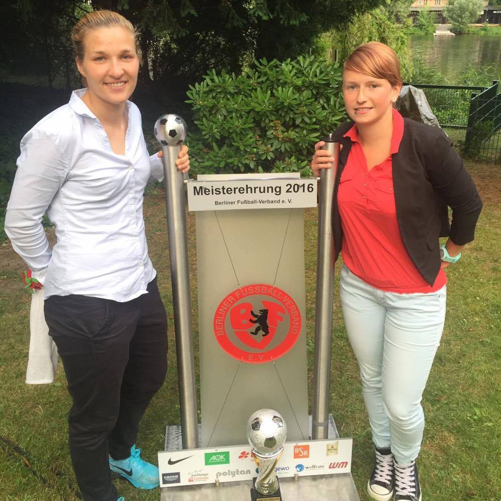 Zwei Frauen posieren neben einer Meisterschaftstafel und einem Pokal, umgeben von grüner Landschaft.