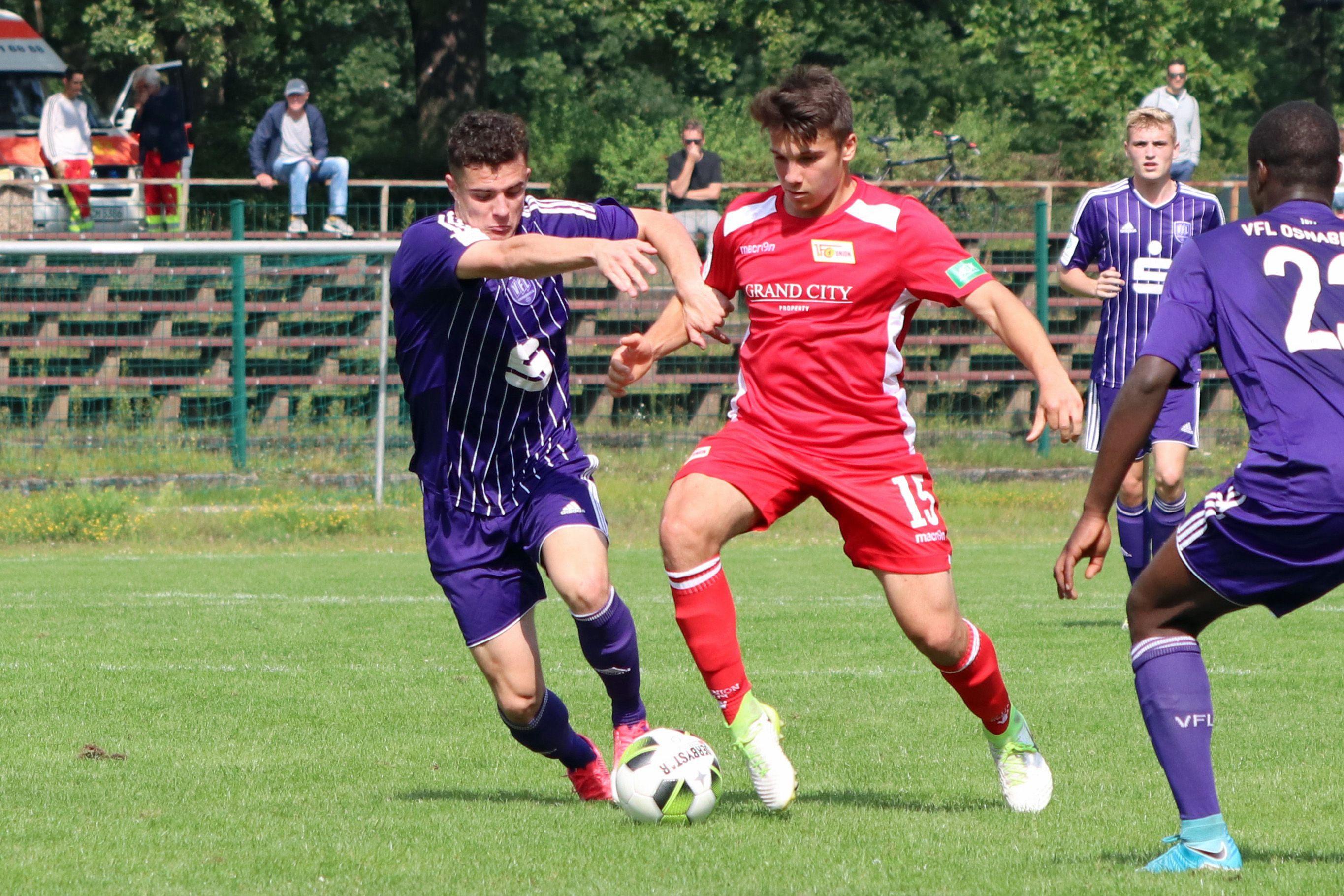 Two soccer players in a duel on a grass field, one in purple and the other in red. Spectators in the background.