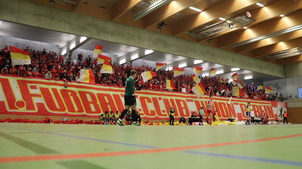 Eine lebhafte Fanszene in einer Halle, mit jubelnden Anhängern und großen Fahnen, die eine Fußballmannschaft unterstützen.