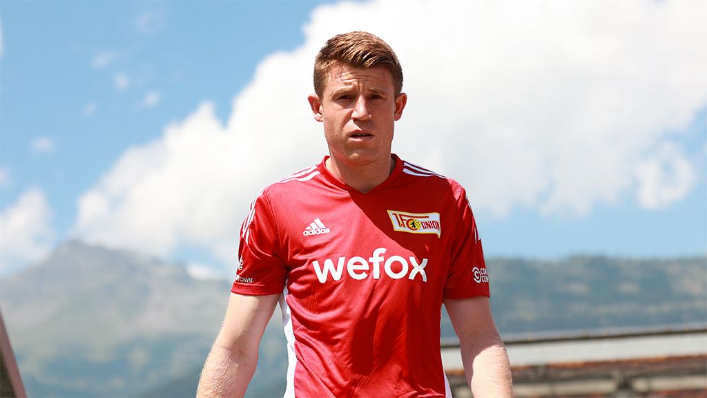 A soccer player in red club gear with the "wefox" logo stands against a blue-white sky.