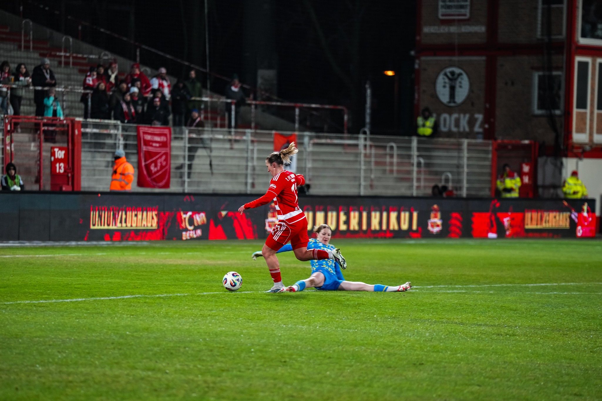Eine Fußballspielszene, in der eine Spielerin im roten Trikot versucht, das Tor zu erreichen, während ein Torwart in blauer Ausrüstung heranrauscht.