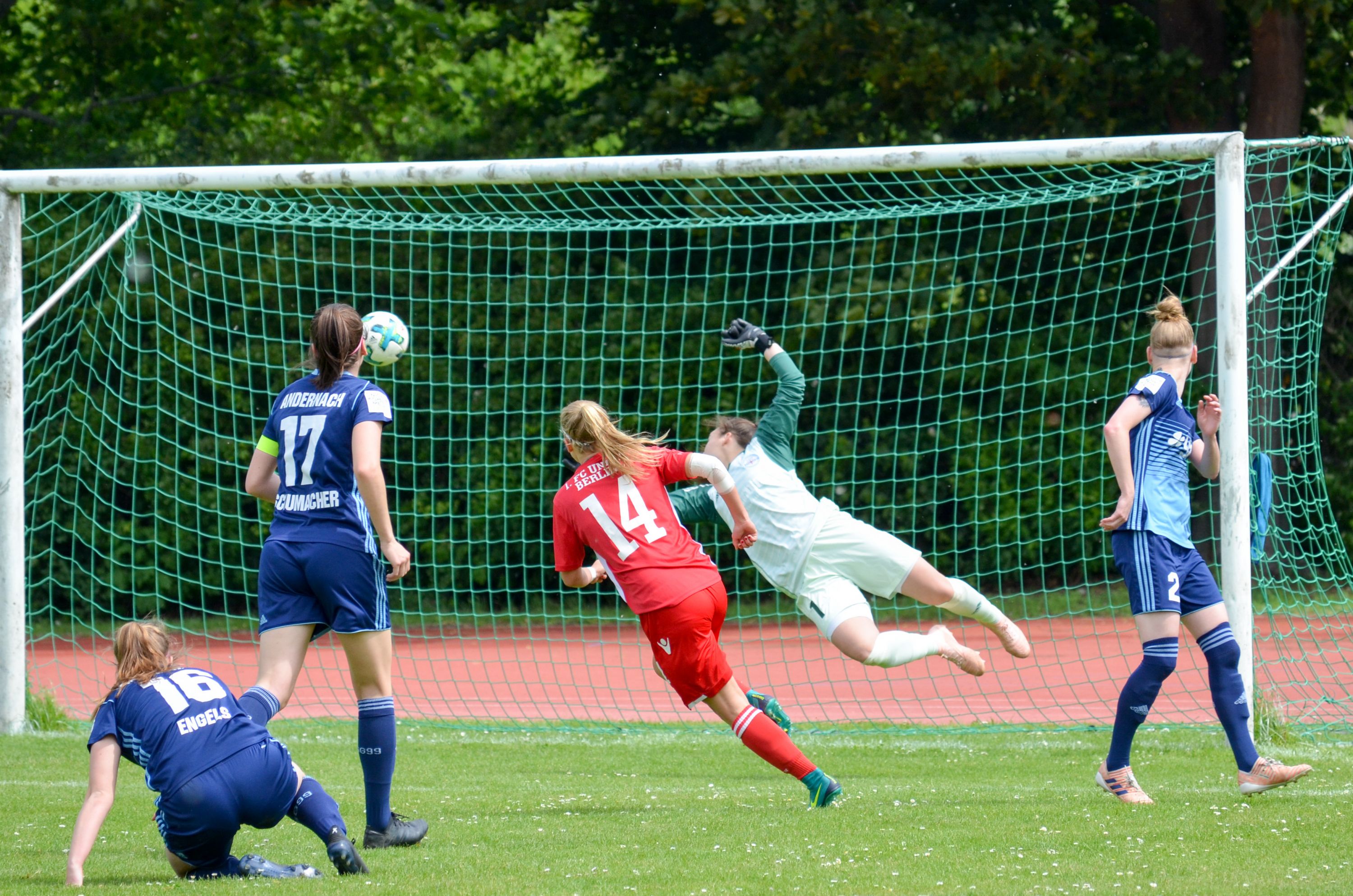 Eine Spielerin in rotem Trikot schießt ein Tor, während die Torhüterin in einem weißen Trikot einen Sprung macht, um den Ball zu halten.