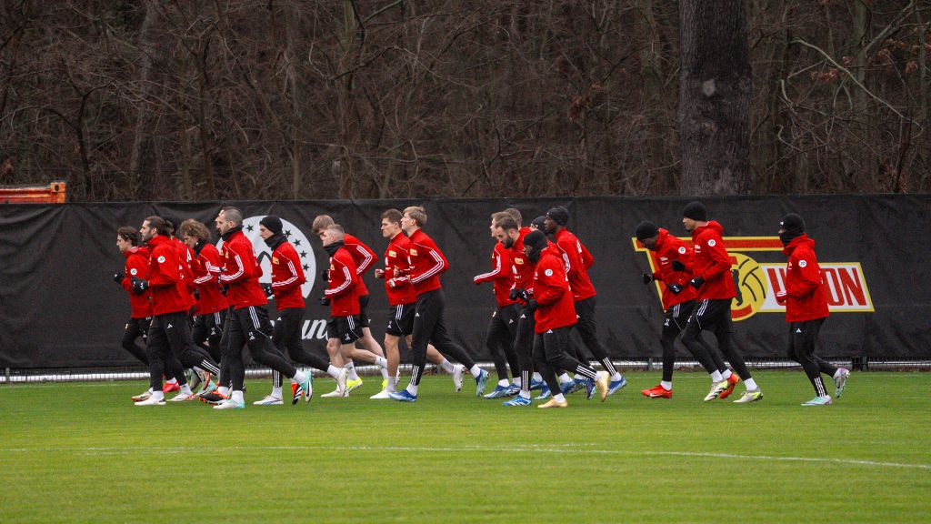 Eine Gruppe von Sportlern in roten Jacken läuft auf einem Trainingsplatz mit grünem Rasen.