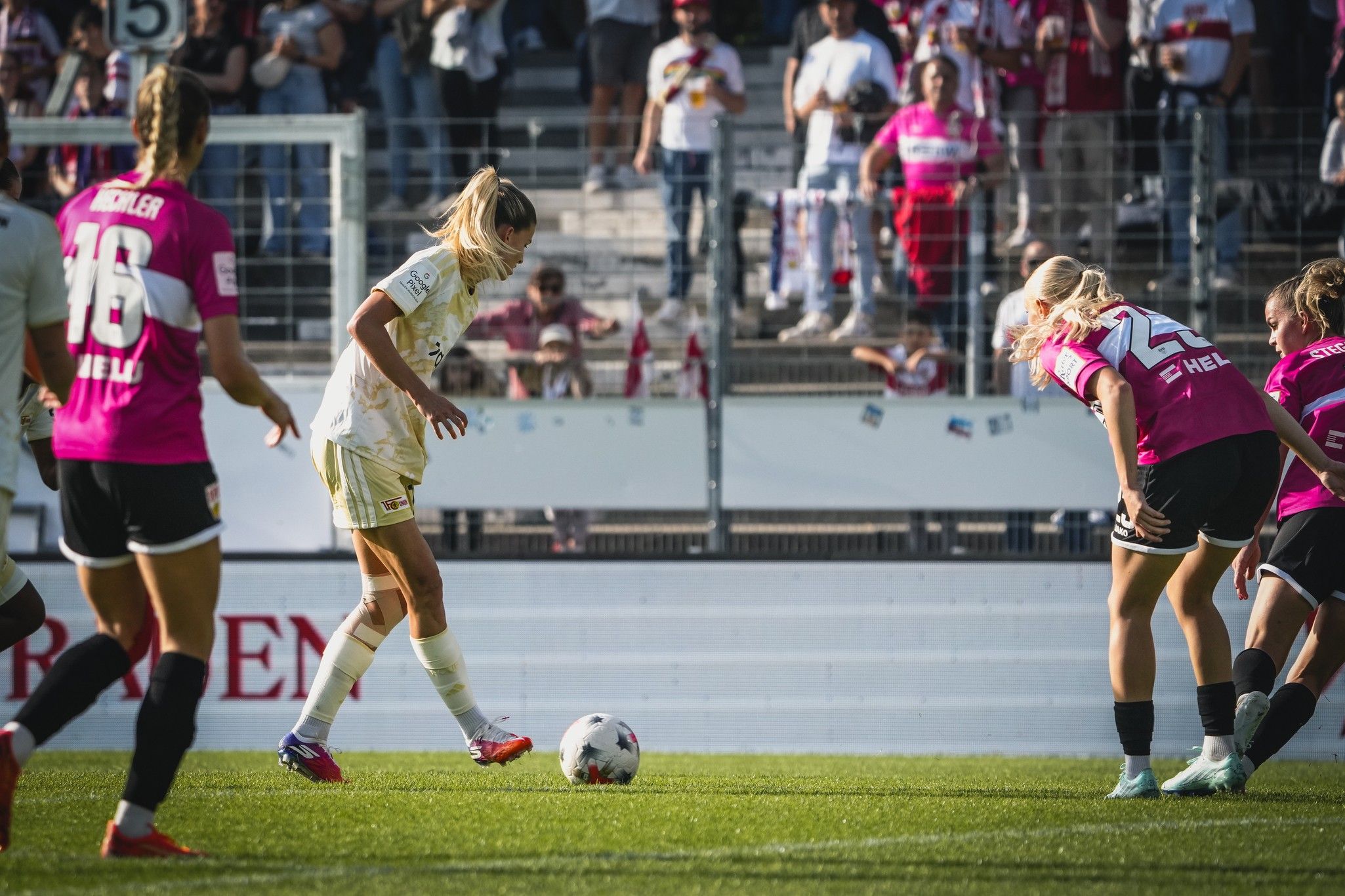 Eine Fußballspielerin in hellen Trikots führt einen Freistoß aus, während Gegnerinnen in pinken Trikots bereitstehen. Zuschauer im Hintergrund.