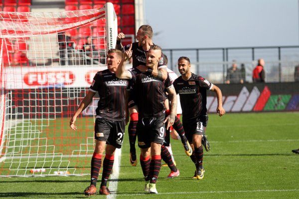 Football players celebrate a goal on the field, with a goal in the background and cheering fans in the stands.