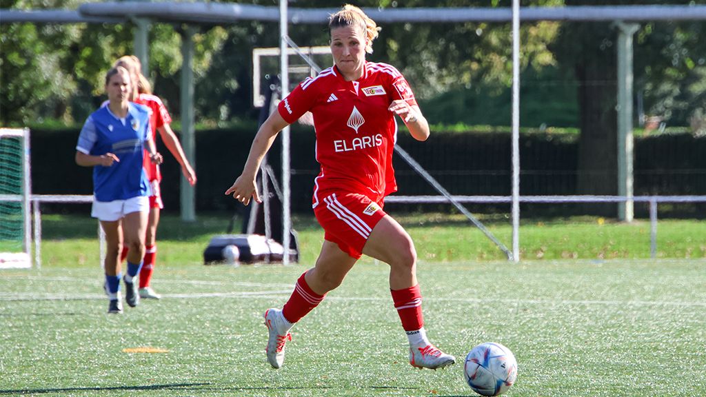 Eine Spielerin in rotem Trikot dribbelt den Ball auf dem Fußballfeld, während eine Gegnerin in Blau im Hintergrund zu sehen ist.