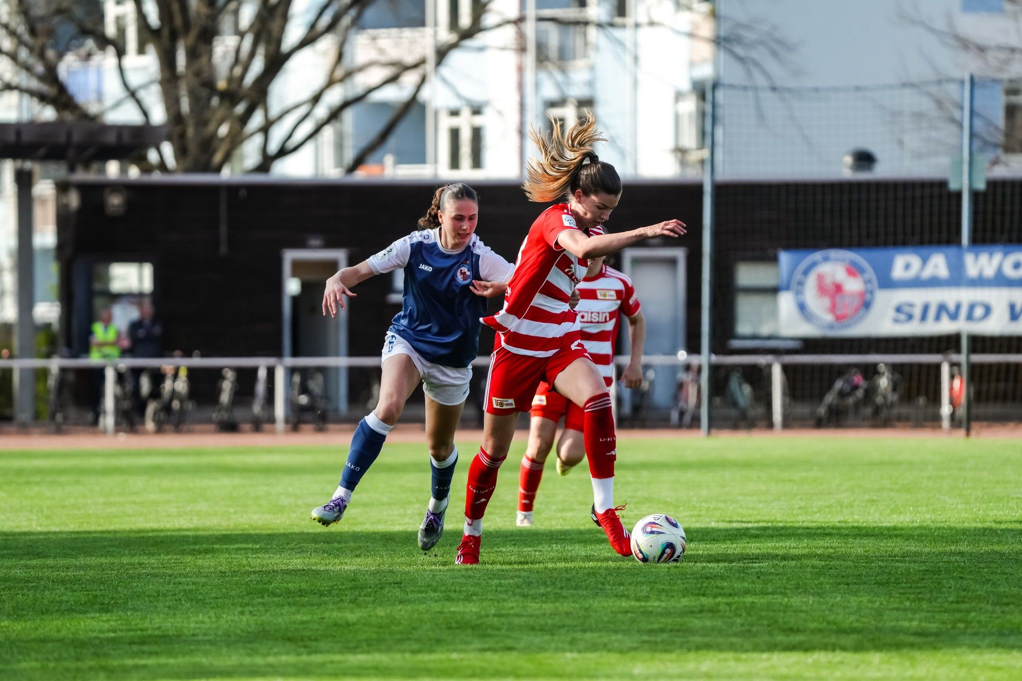 Zwei Fußballspielerinnen im Wettkampf auf dem Feld; eine in roter, die andere in blauer Uniform.