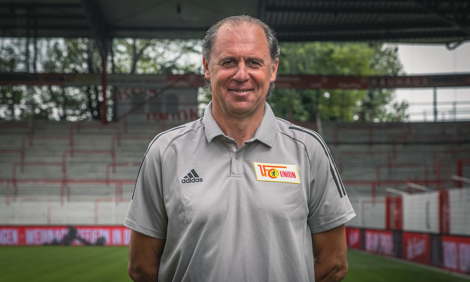 Ein lächelnder Mann in einem grauen Sportpoloshirt steht im Stadion, im Hintergrund sind leere Tribünen zu sehen.