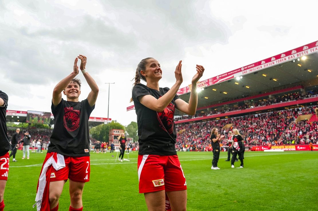 Zwei Fußballspielerinnen in roten Trikots applaudieren jubelnd auf dem Spielfeld vor einer Menge in einem Stadion.