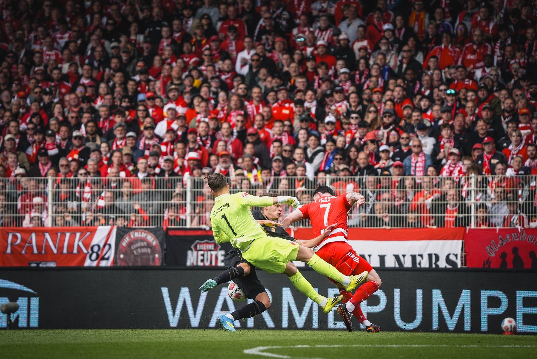 Ein Fußballspieler fällt nach einem Zweikampf im Stadion, im Hintergrund jubelnde Fans mit Vereinsfarben.