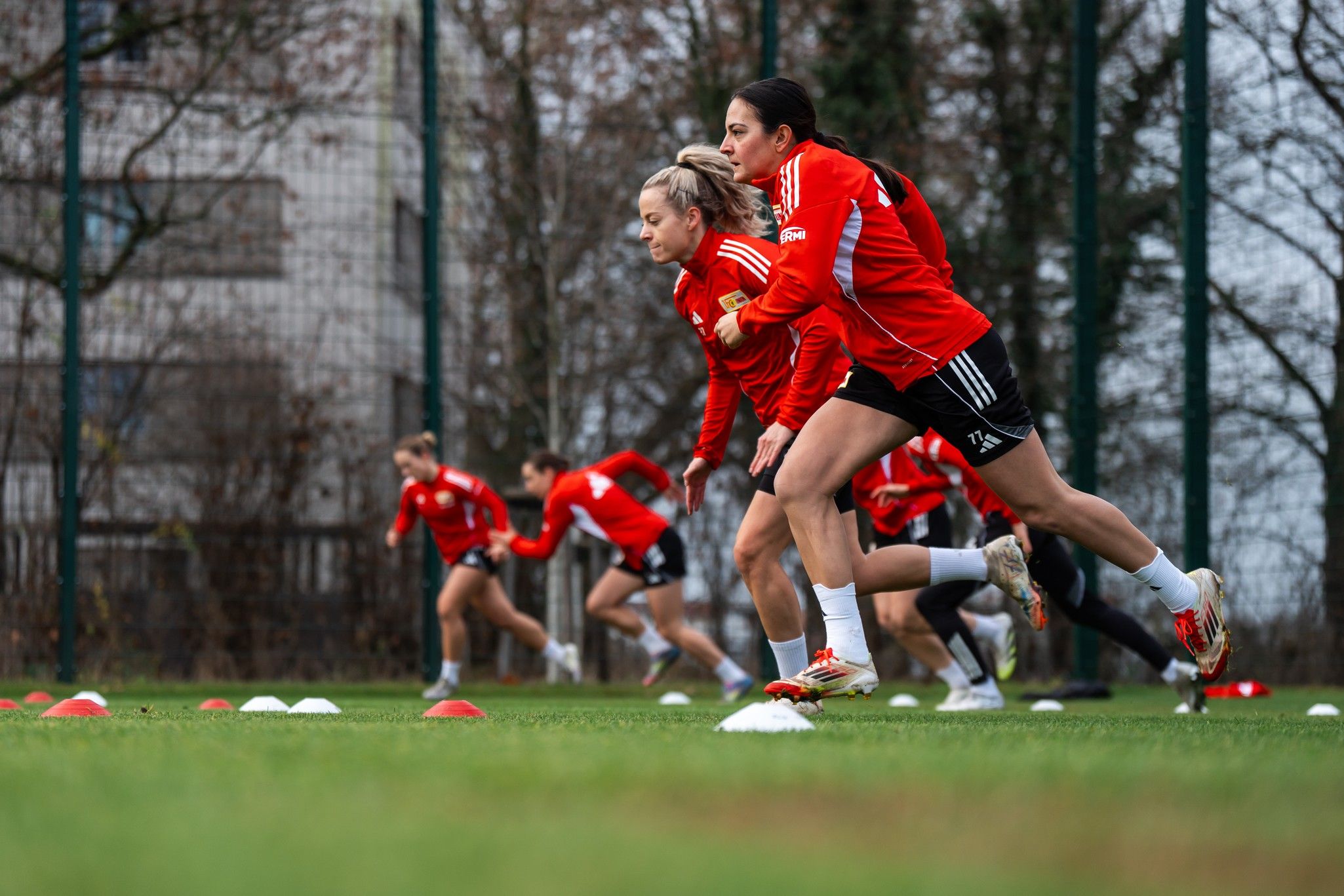 Zwei weibliche Fußballspielerinnen sprinten im Training, während weitere Spielerinnen im Hintergrund Übungen durchführen.