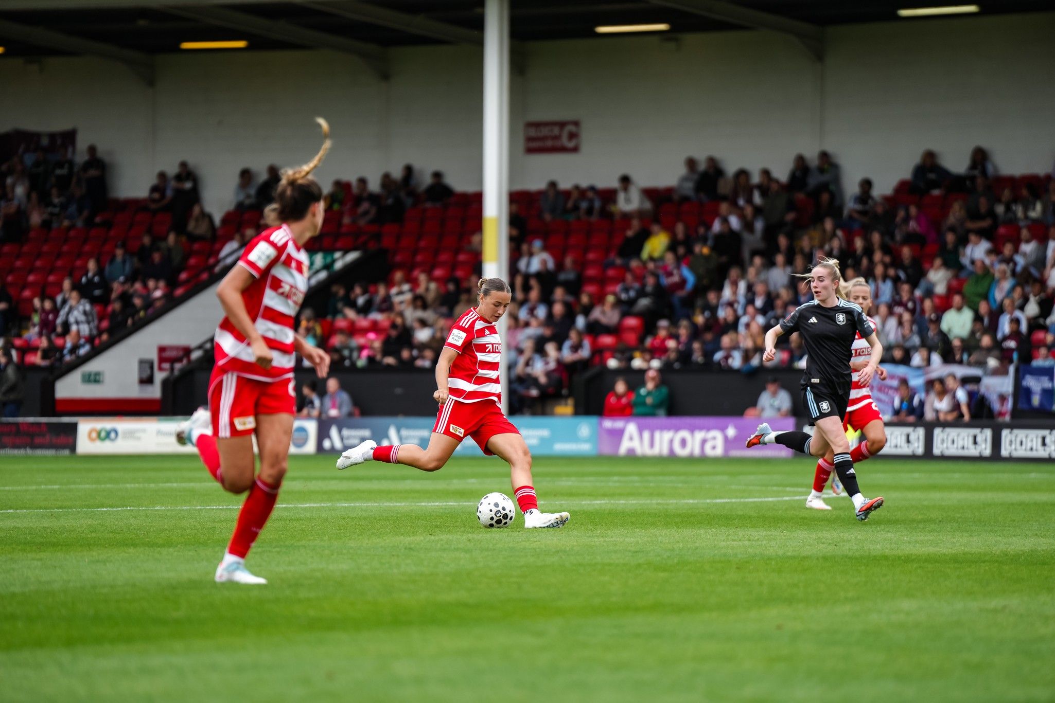 Spielszene aus einem Fußballspiel, bei dem zwei Teams auf dem Rasen agieren und eine Spielerin den Ball führt. Publikum im Hintergrund.
