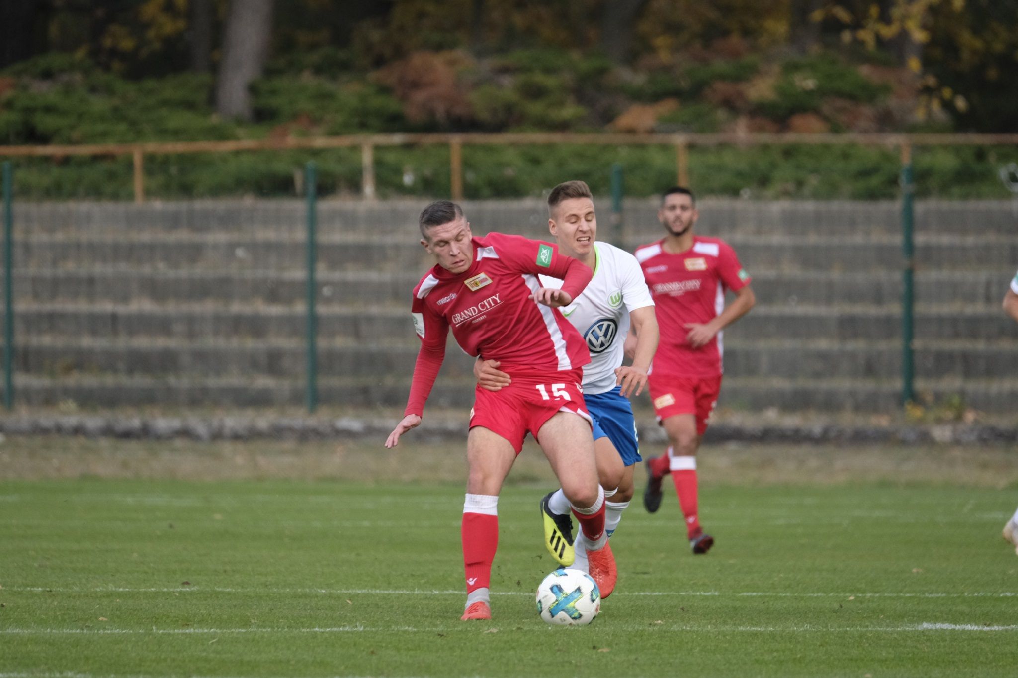 Ein Fußballspiel, bei dem ein Spieler in Rot einen anderen Spieler in Blau herausfordert, während ein weiterer Spieler im Hintergrund zuschaut.