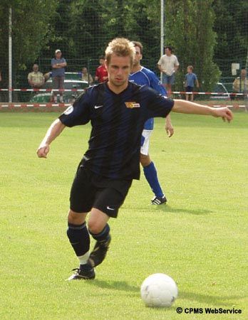 Ein Fußballspieler in schwarzer Uniform tritt einen Ball auf einem Rasenplatz, während ein anderer Spieler im Hintergrund steht.