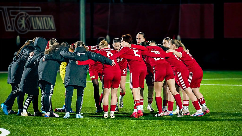 Frauenfußballteam in roten Trikots bildet einen Kreis auf dem Spielfeld und bereitet sich auf das Spiel vor.