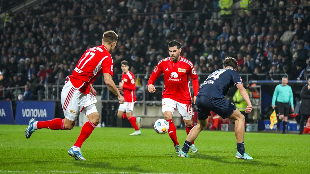 Zwei Fußballspieler in roten Trikots verfolgen einen dritten Spieler in dunkelblauem Trikot auf dem Spielfeld. Zuschauer im Hintergrund.
