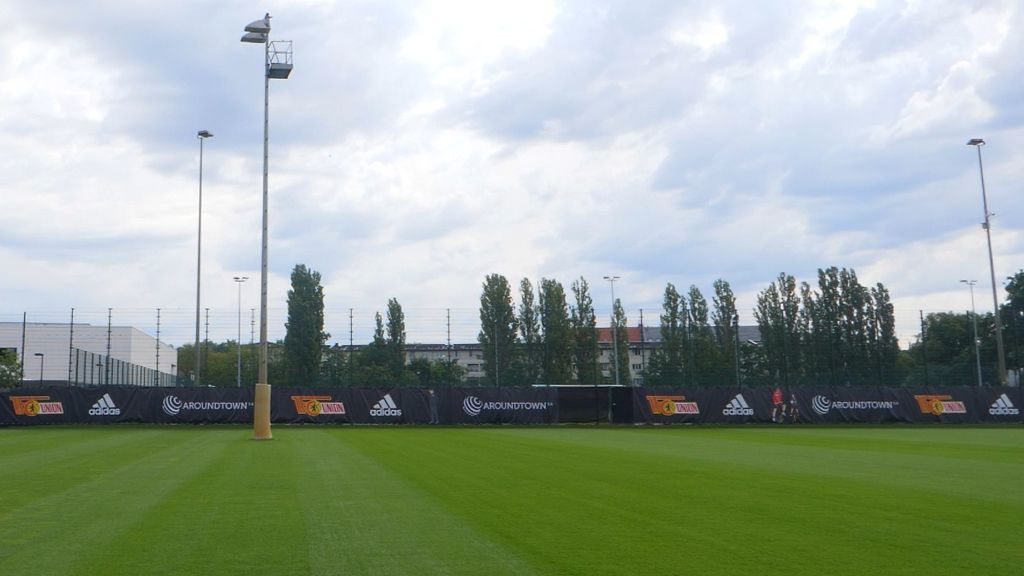 Green football field with advertising banners and several trees in the background under a cloudy sky.