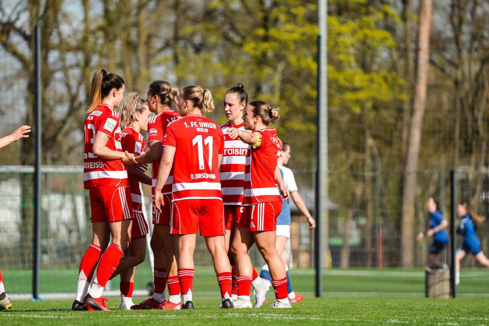 Spielerinnen eines Fußballteams in roten Trikots feiern ein Tor auf dem Spielfeld. Bäume und Zuschauer im Hintergrund.