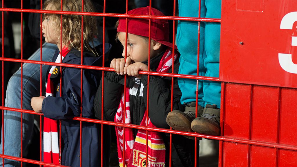 Ein Kind in einem roten Hut und Schal lehnt mit beiden Händen an einem Gitter, während es einen Blick aus dem Stadion wirft.