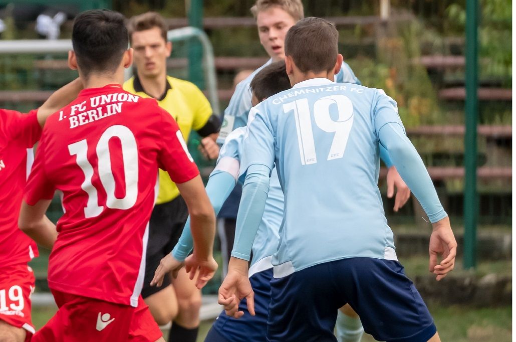 Two soccer players in blue and red uniforms face each other on a field while a referee watches.