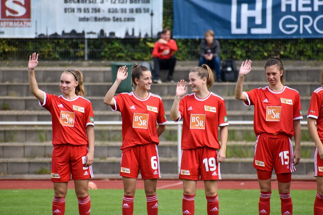 Fünf Fußballspielerinnen in roten Trikots winken lächelnd zum Publikum.