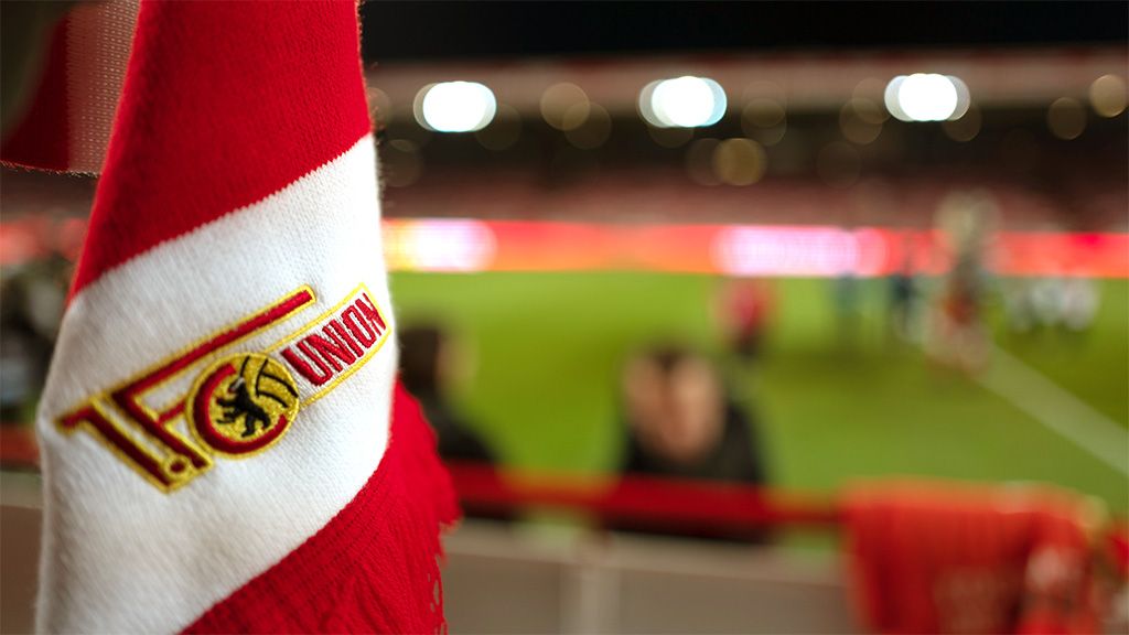 Fokus auf das Vereinswappen von Union Berlin auf einem roten Schal im Vordergrund, Unschärfe der Zuschauer und des Spielfelds im Hintergrund.