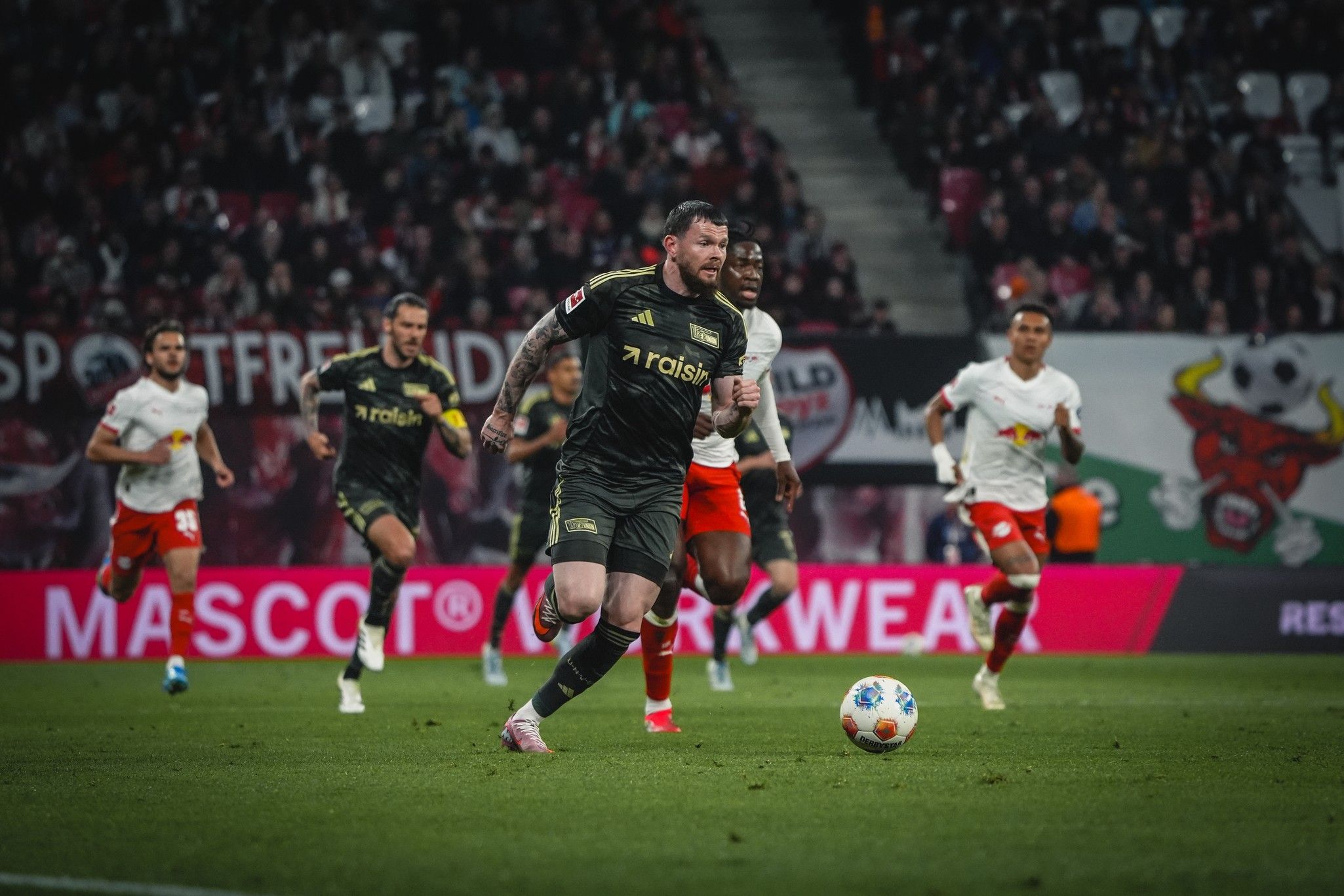 Ein Fußballspieler in schwarzer Ausstattung jagt den Ball, während weitere Spieler im Hintergrund agieren und Zuschauer im Stadion zu sehen sind.