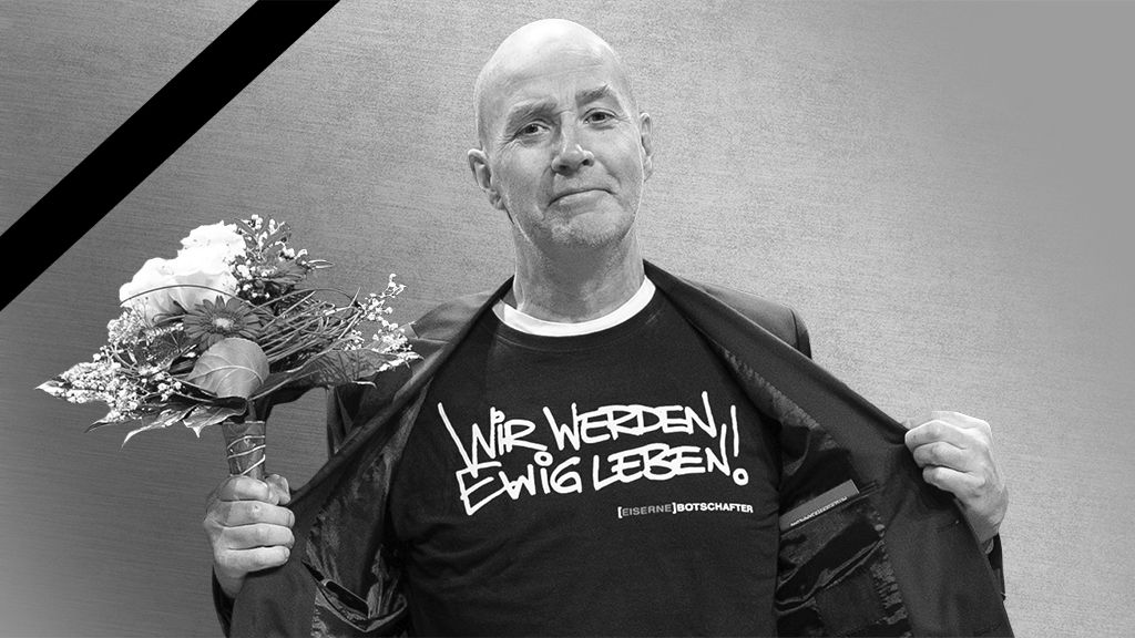 Ein Mann in einem schwarzen Shirt mit dem Aufdruck „Wir werden ewig leben!“ hält Blumen in der Hand und hat einen feierlichen Gesichtsausdruck.
